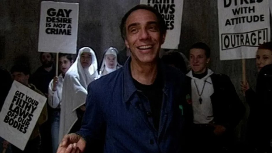 Smiling man facing camera with a group of people in nun costumes and Outrage placards behind 
