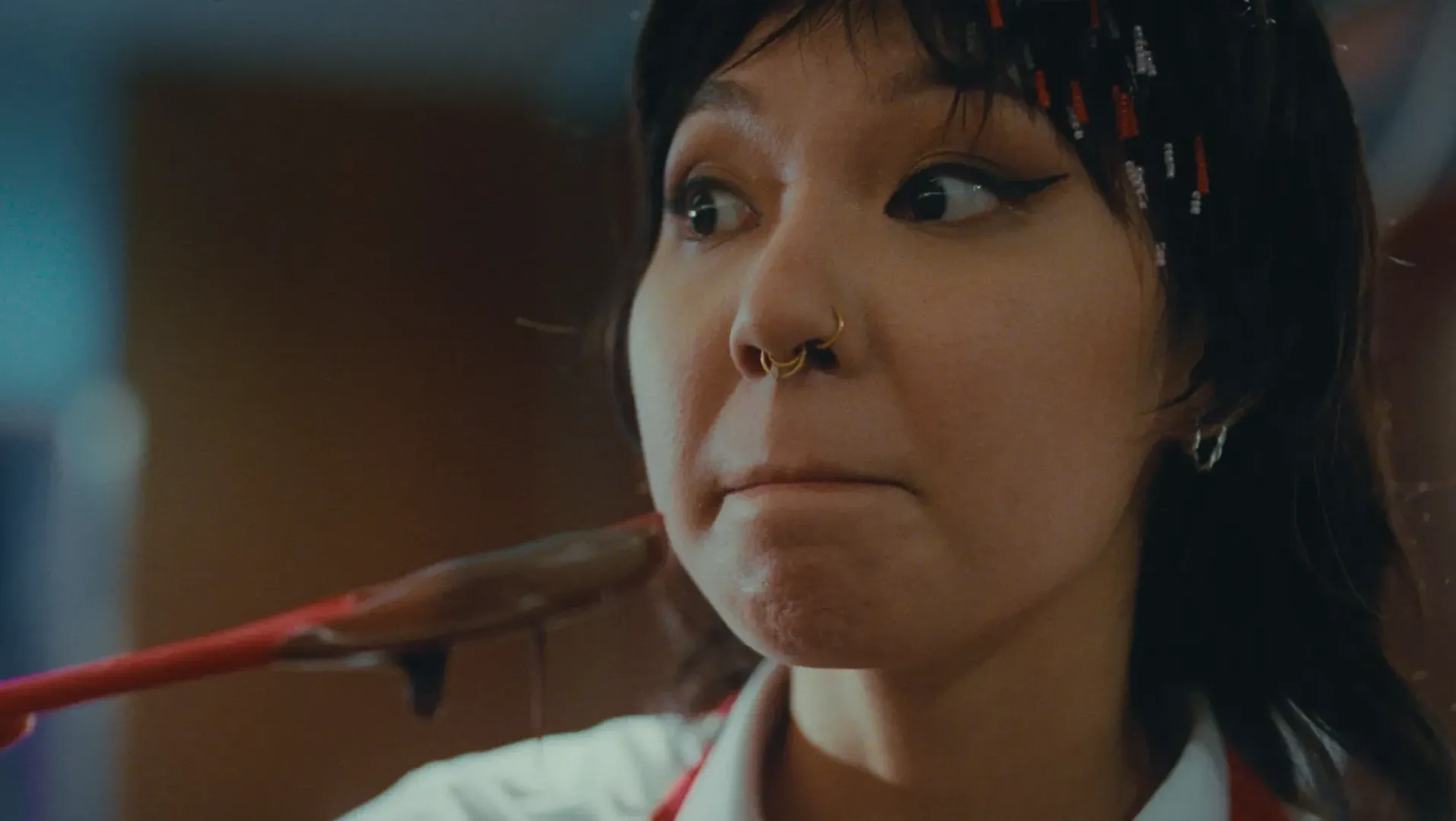Close-up: a woman pauses mid-taste, chocolate dripping from a red spatula still at her lips.