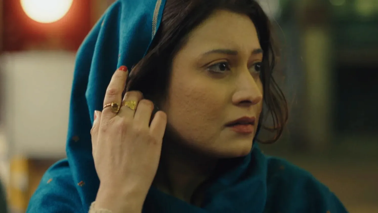 Woman in blue headscarf looks to the side with one hand holding her scarf away from her face 