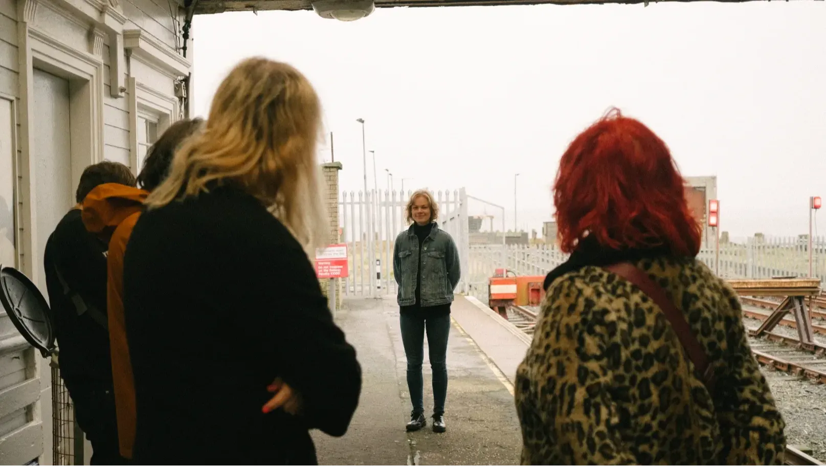 4 people are seen from behind standing in a semi circle facing a woman in a denim jacket  on a train station platform