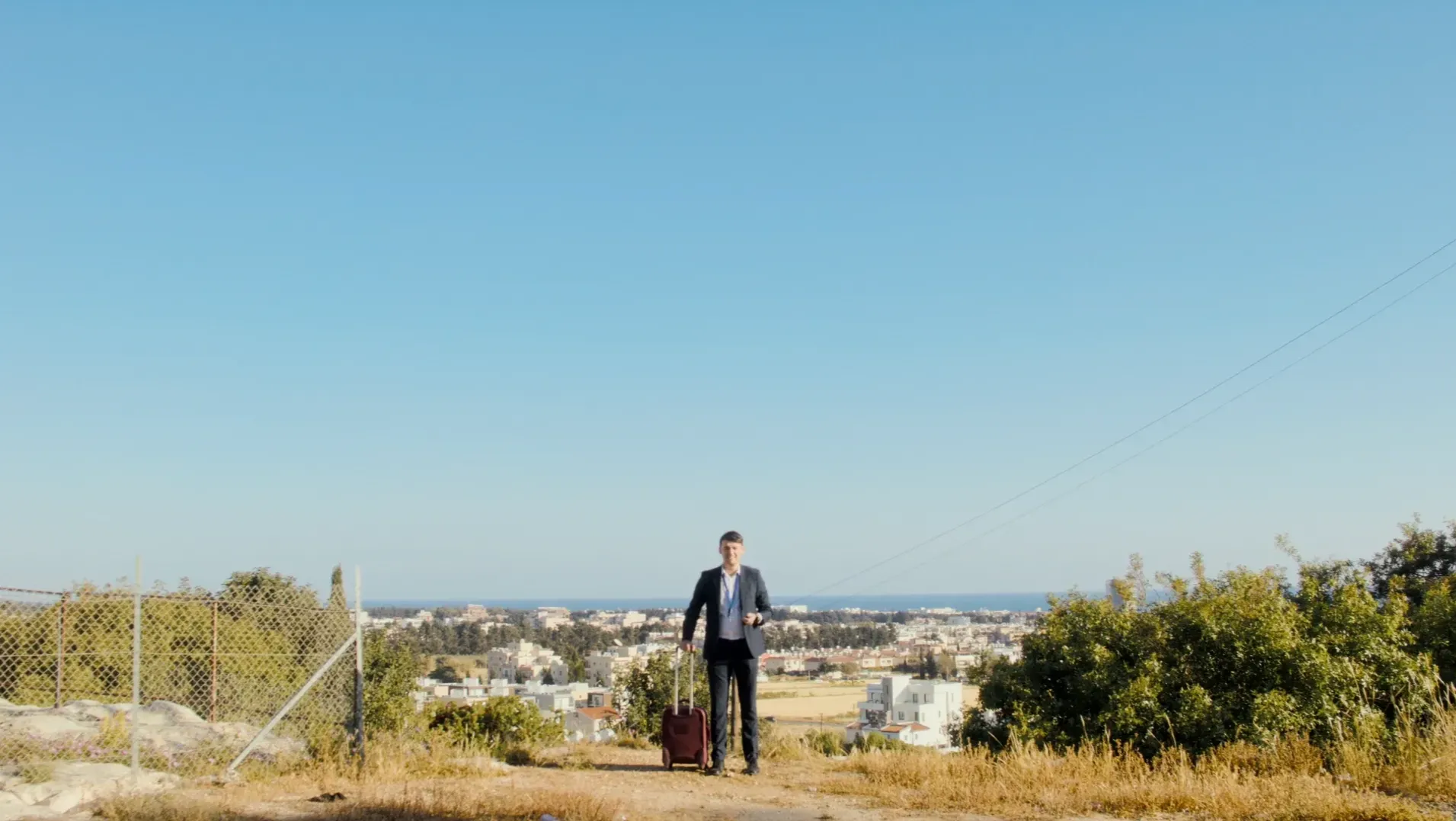 'Bill' an awkward-looking business man dressed in a suit with a wheelie travel case waits in the middle of a rusty path, cypriot landscape, a large blue sky above and the sea in the distance.