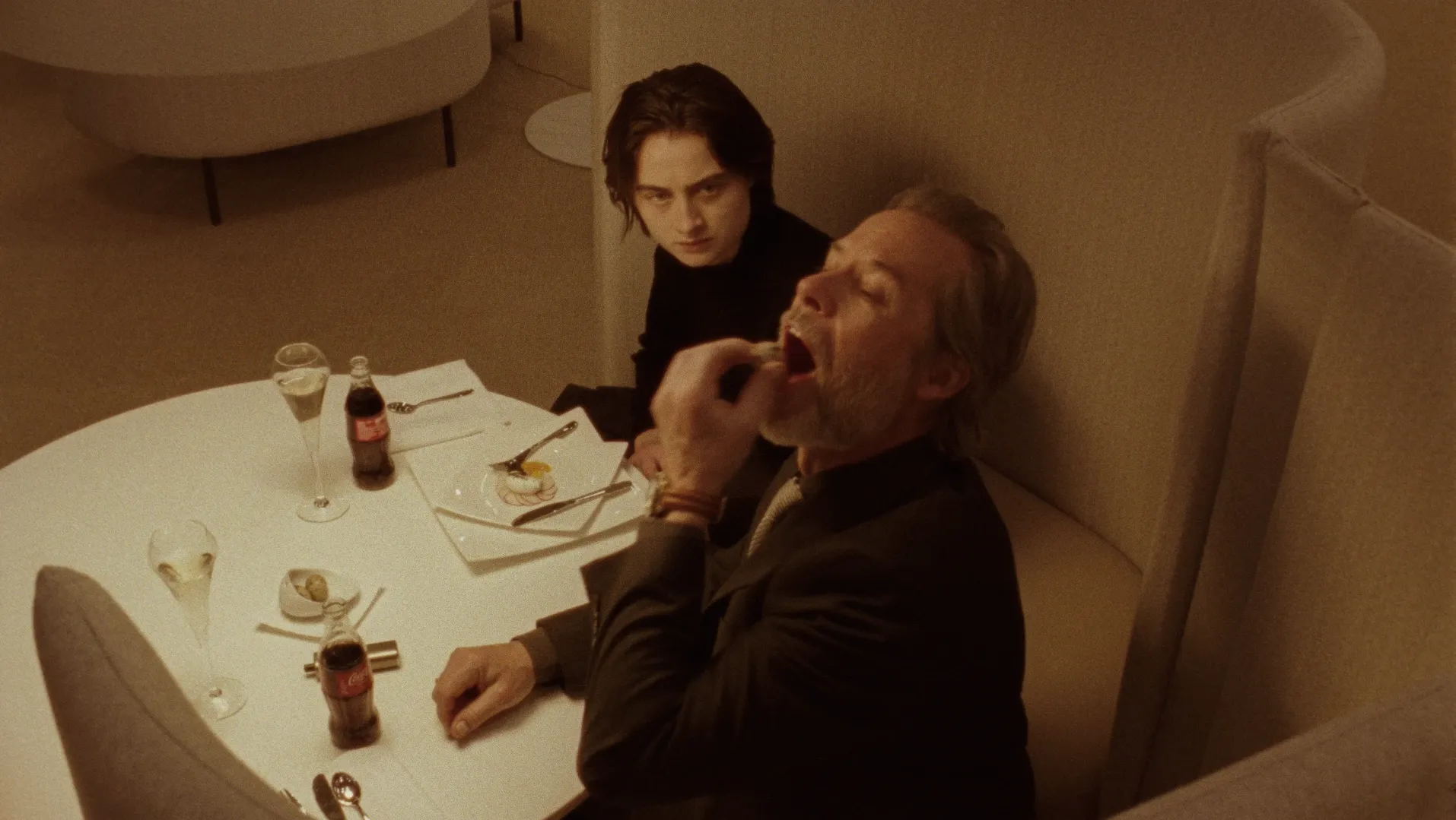 A young person is looking at man who is putting a quail egg in his mouth. They are both sitting down at a table in an airport lounge.