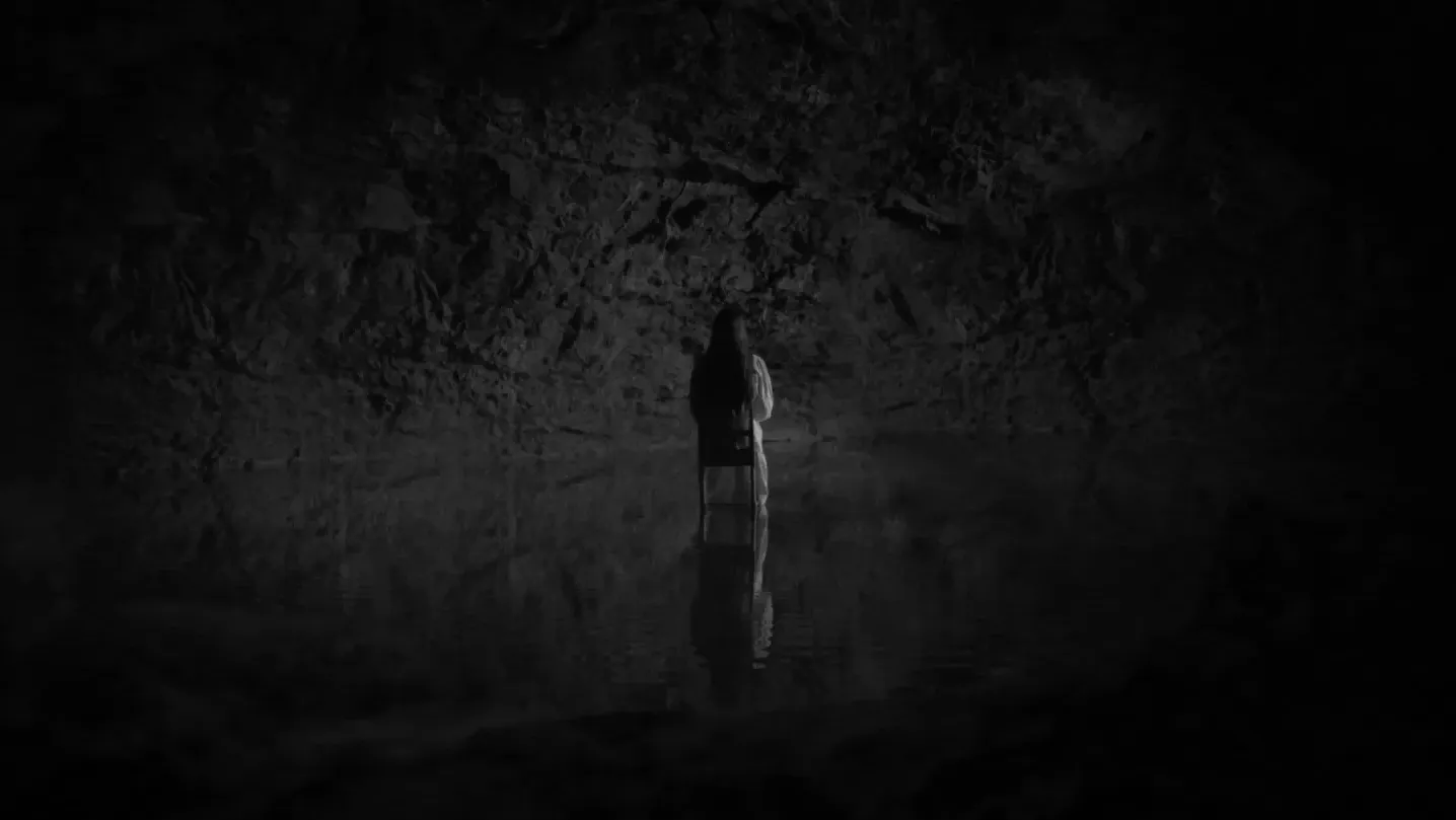 A black white image in a cave of a woman sitting on a chair that seeming floats on dark water