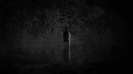 A black white image in a cave of a woman sitting on a chair that seeming floats on dark water