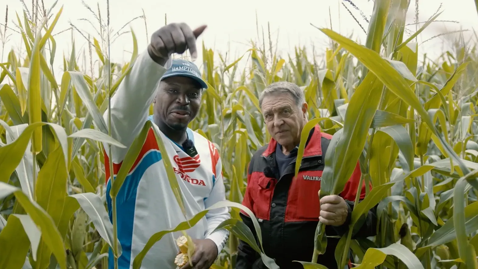 Fekky, in a white sports jumper and blue cap, smiles and points down beside Charlie in red farmer gear. They stand in tall cornfields, with Fekky holding sweetco