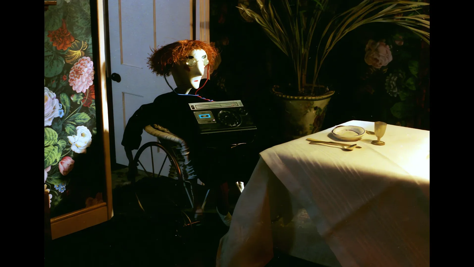 A red-haired puppet in round glasses sits in a wheelchair with a radio on its lap beside a laid table