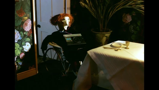 A red-haired puppet in round glasses sits in a wheelchair with a radio on its lap beside a laid table