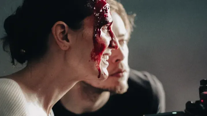 A white woman with fake blood on her face looks into a camera playback screen. A white man looks as well.
