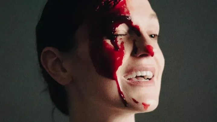 A white woman's face, very contrasted with light and shadow. She has fake blood all over one half of her face and nose. She is smiling and has one eye closed.