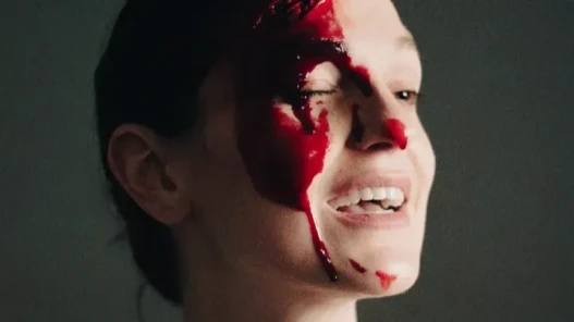A white woman's face, very contrasted with light and shadow. She has fake blood all over one half of her face and nose. She is smiling and has one eye closed.