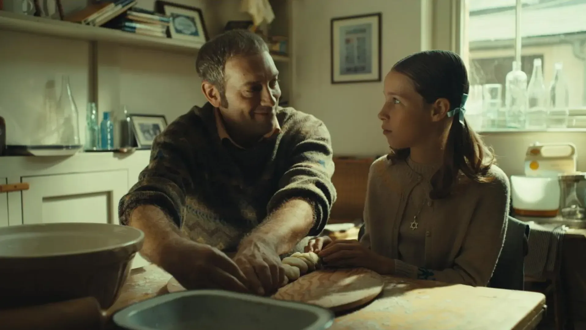 A man and a young girl sit at a kitchen table, preparing braided dough together. The man smiles at the girl, who looks back at him. Warm sunlight streams through the window.