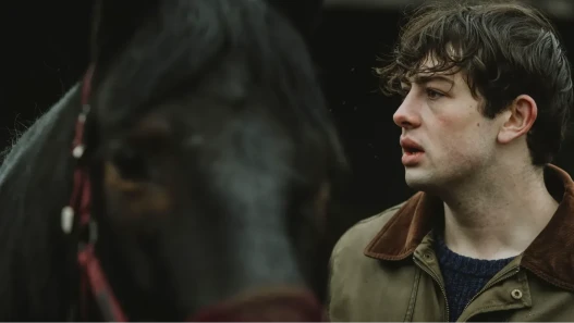 Close-up of a young man with wearing a green jacket, looking anxiously off-screen, with a black horse in the foreground partially obscuring his face.