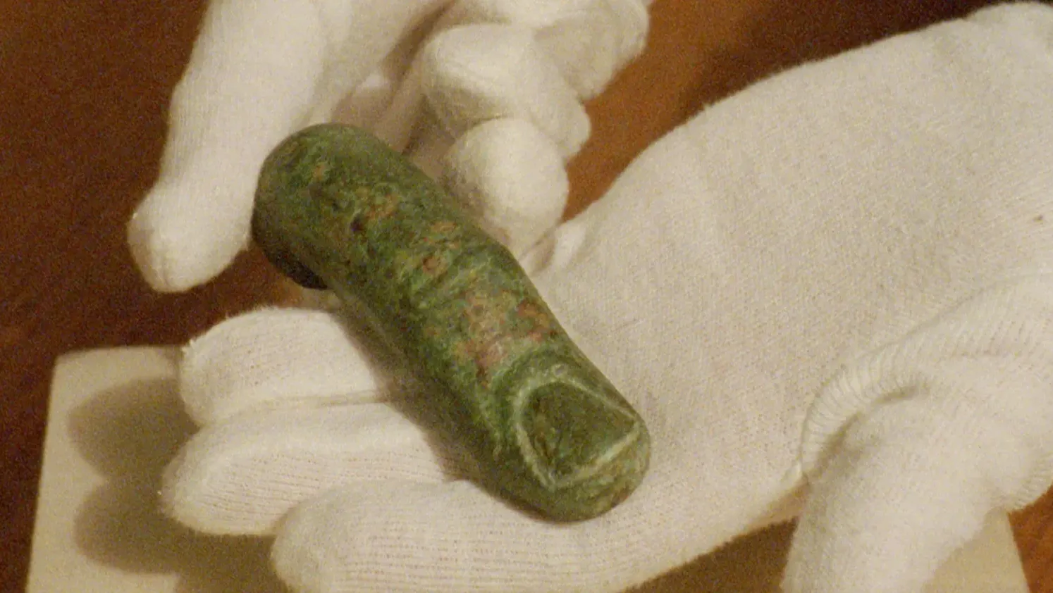 Close-up of white-gloved hands holding a small, weathered bronze Roman artefact with a green patina, captured on grainy 16mm film.