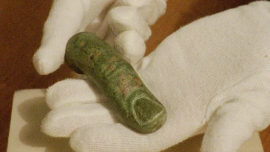 Close-up of white-gloved hands holding a small, weathered bronze Roman artefact with a green patina, captured on grainy 16mm film.