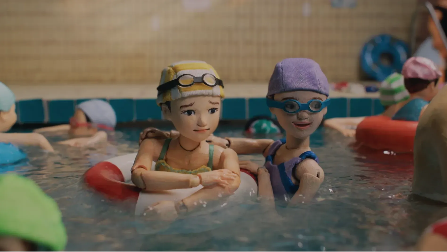 Two girls floating in a swimming pool, one wearing a swim ring, both looking towards the deep end